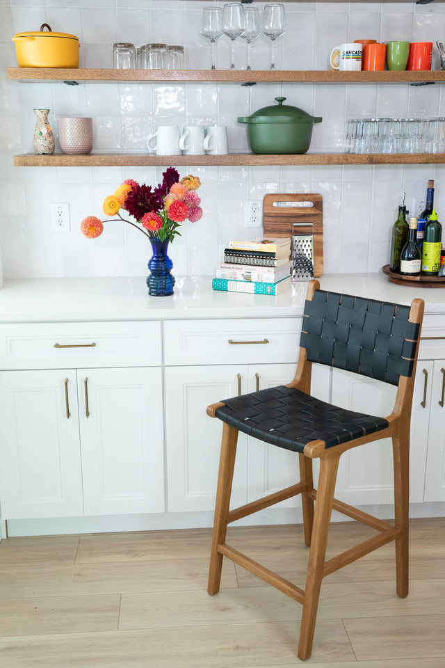 A wooden counter stool with a woven black leather seat and back, set against white cabinets and open shelves holding colorful dishes and a bright bouquet in a blue vase.