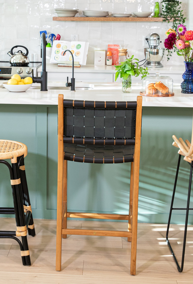 A wooden counter stool with a woven black leather back, positioned at a teal kitchen island with a black faucet and colorful decor in the background.
