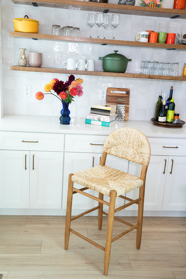 A wooden counter stool with a rounded woven back and seat set against white cabinets, open shelves of colorful dishes, and a bright floral arrangement in a blue vase.