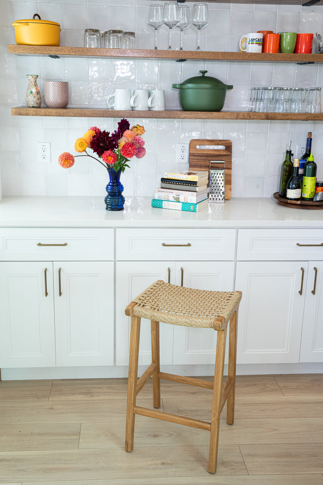 Wooden counter stool with a woven seat in front of white cabinets, open shelves of colorful dishes, and a blue vase filled with bright flowers.