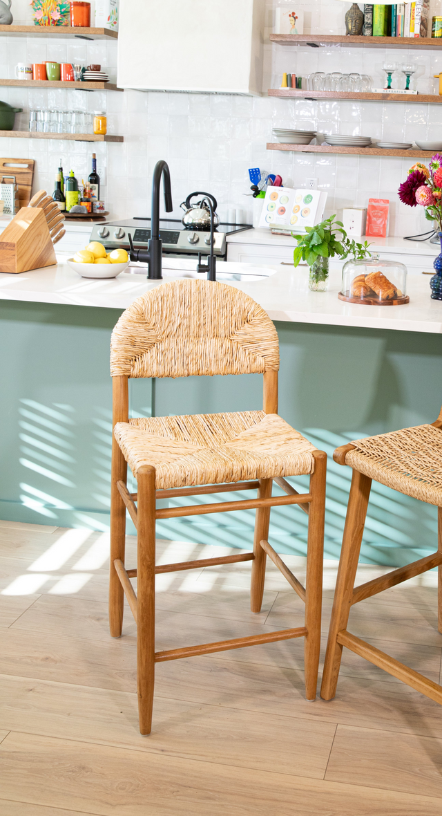A wooden counter stool with a rounded woven back and seat set against a teal kitchen island, black faucet, and bright decor accents in the background.