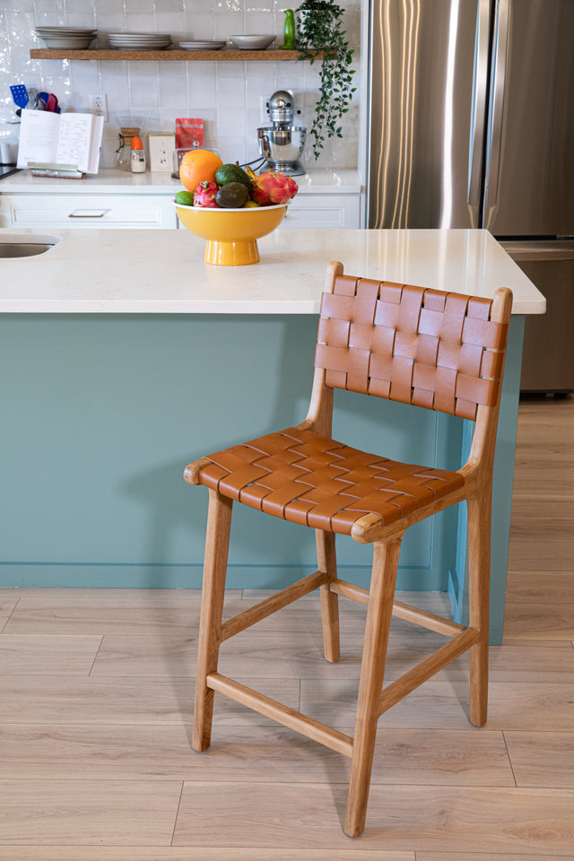 A wooden counter stool with a woven warm brown leather seat and back, positioned beside a teal kitchen island topped with a bowl of colorful fruit