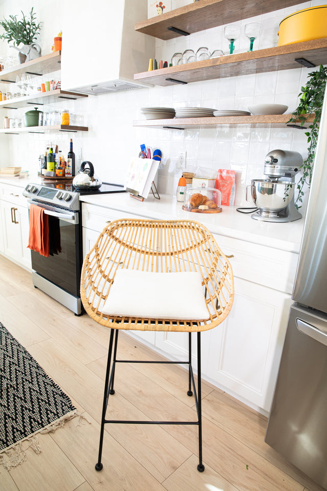 A rattan counter stool with a curved woven seat, white cushion, and black metal legs, positioned in a bright kitchen with open shelving, white cabinetry, and modern appliances.