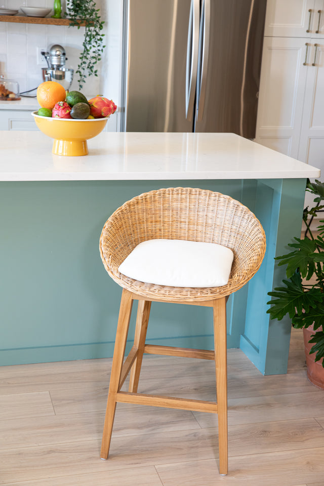 A wooden counter stool with a rounded wicker seat, a white cushion, and a teal kitchen island topped with fresh fruit in the background.