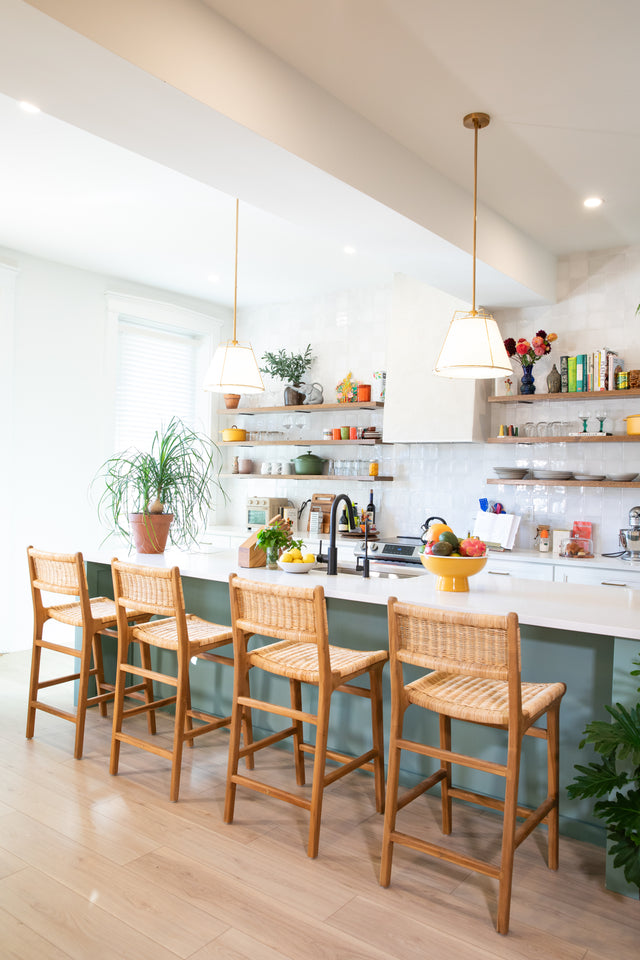 Four wooden counter stools with woven rattan seats lining a teal kitchen island, surrounded by open shelves, pendant lights, and vibrant potted plants