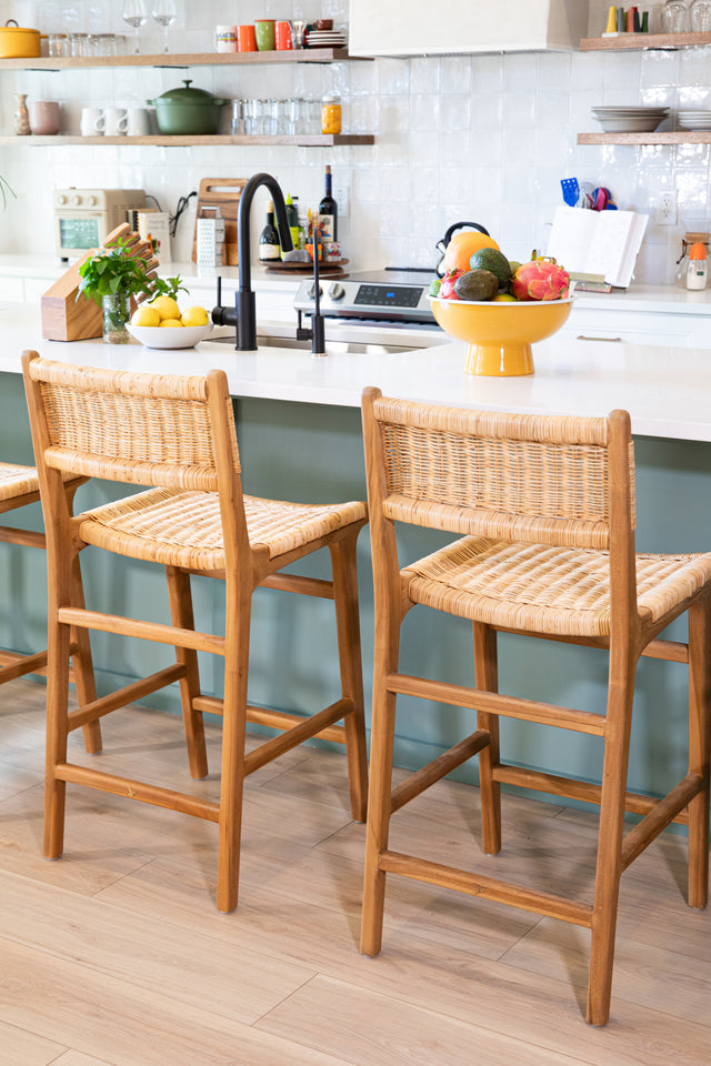 Wooden counter stools with woven rattan seats, placed at a teal kitchen island featuring a black faucet, a colorful fruit bowl, and open shelving in the background.