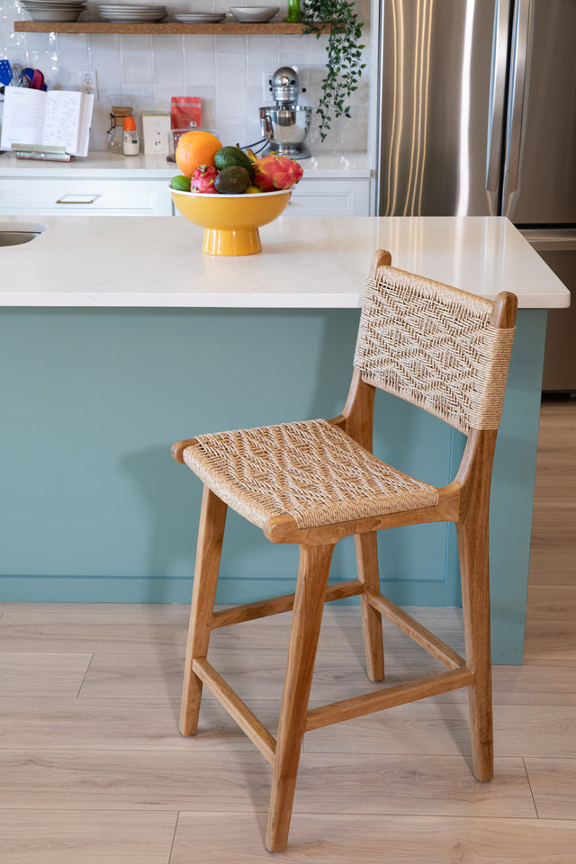 A wooden counter stool with a detailed woven seat and back in a geometric pattern, positioned beside a teal kitchen island topped with a bowl of fresh fruit.