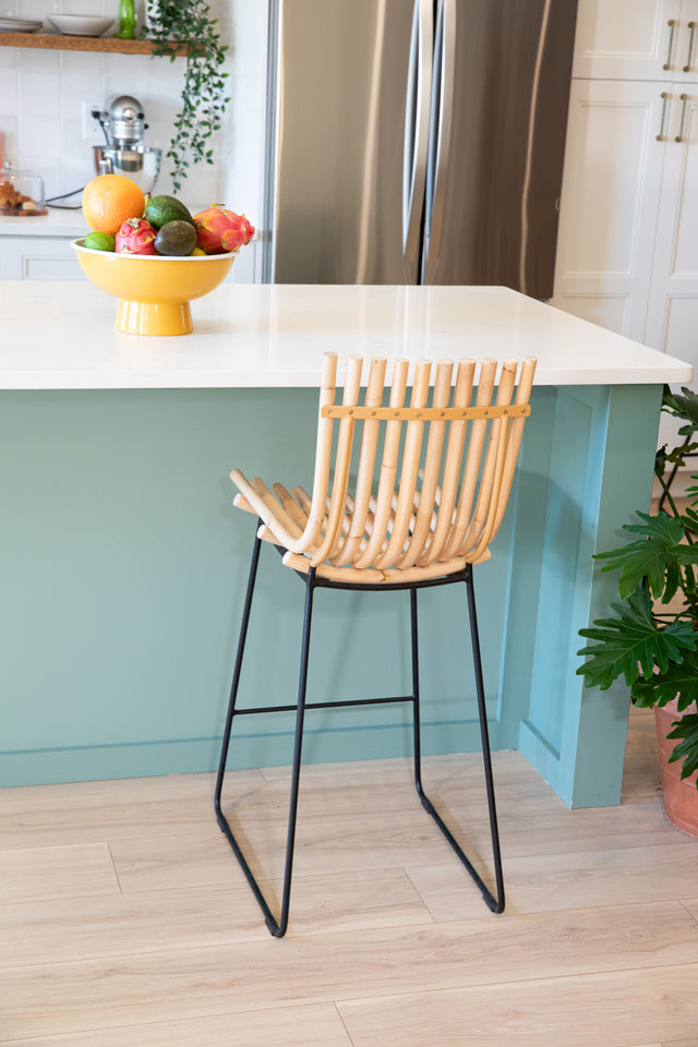 Modern counter stool with curved wooden slats and black metal legs next to a teal kitchen island topped with a colorful fruit bowl.