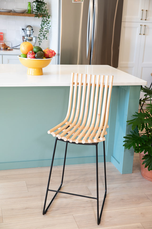 Modern counter stool with curved wooden slats and black metal legs next to a teal kitchen island topped with a colorful fruit bowl.