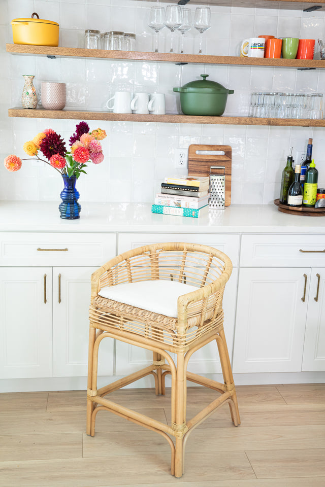 A rattan counter stool with a rounded back, white cushion, and sturdy woven frame, set against white cabinets and open shelves with colorful dishes and a vase of vibrant flowers