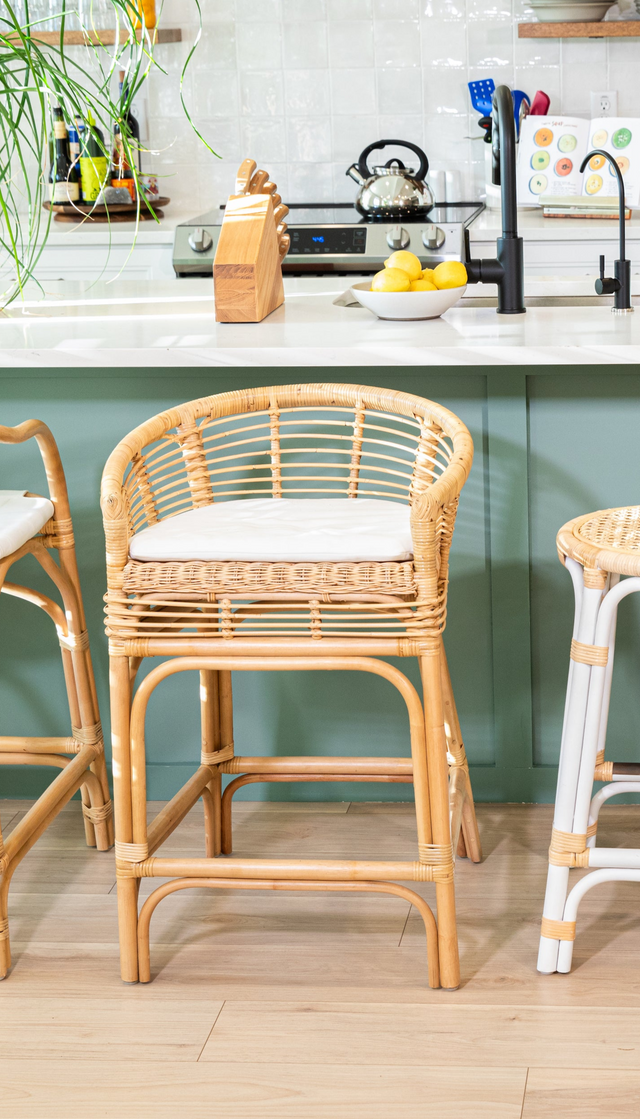 A rattan counter stool with a rounded back, white cushion, and curved details, positioned at a teal kitchen island with a black faucet and modern decor.