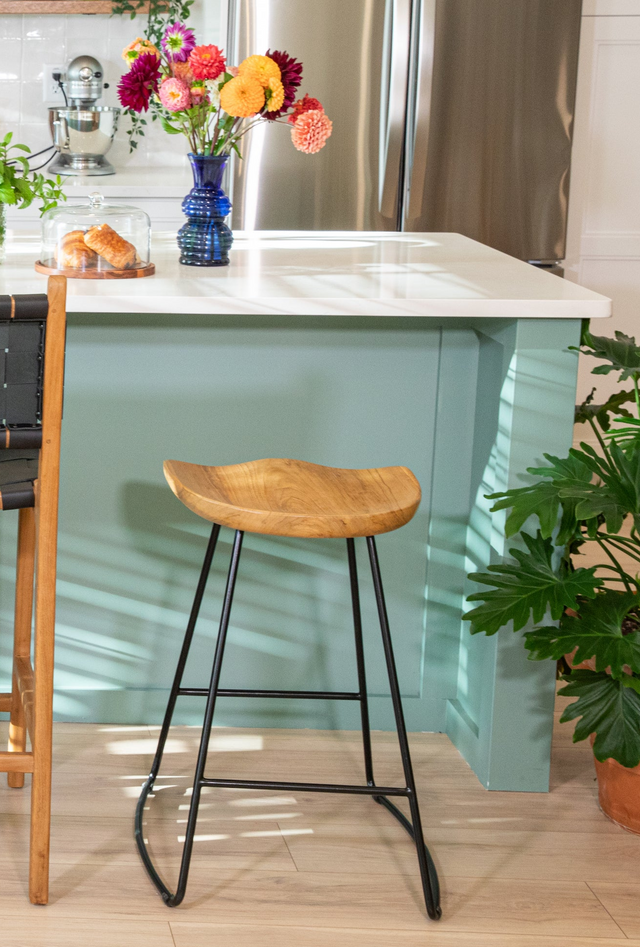 A counter stool featuring a sculpted wooden seat and minimalist black metal legs, placed at a teal kitchen island adorned with colorful flowers in a vase and baked goods