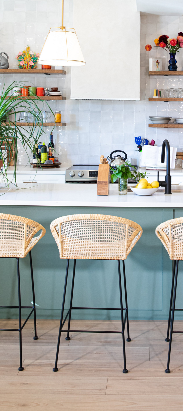 Three rattan counter stools with curved woven seats and black metal legs at a teal kitchen island, with open shelving, potted plants, and a modern black faucet in the background.