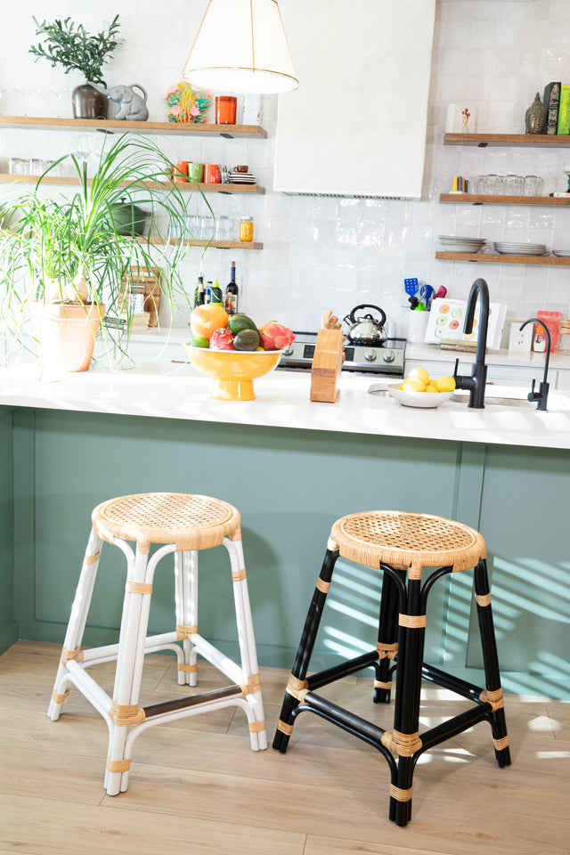 Two rattan counter stools, one with a black frame and the other with a white frame, featuring round woven seats, positioned at a teal kitchen island with potted plants and vibrant decor