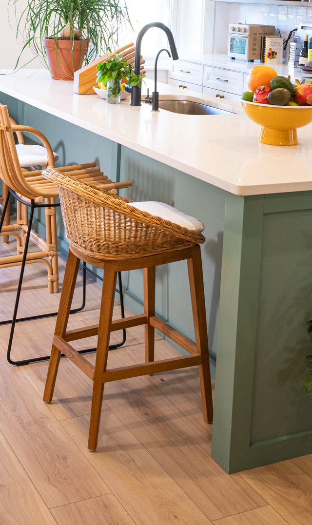 A wooden counter stool with a woven, basket-like seat and cushion, positioned against a teal kitchen island with a black faucet and bowl of colorful fruits.