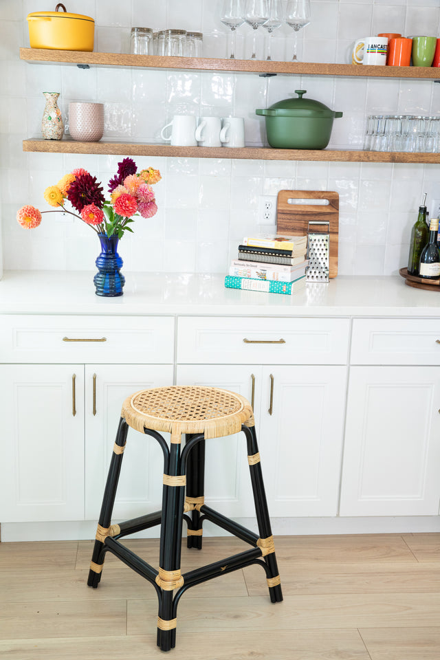 A black-framed rattan counter stool with a round woven seat, placed in front of white cabinets and open shelving decorated with colorful dishes and fresh flowers in a vase.