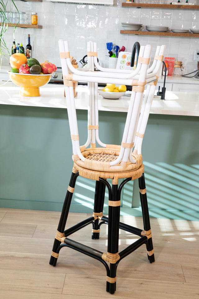 A black-framed rattan counter stool with a stacked white-framed stool on top, showcasing round woven seats, positioned by a teal kitchen island in a bright and modern kitchen.