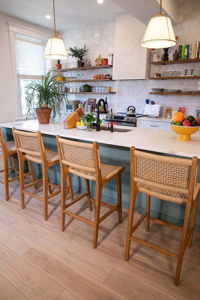 Four wooden counter stools with woven seats lined up at a white kitchen island, surrounded by open shelves, potted plants, and pendant lights.