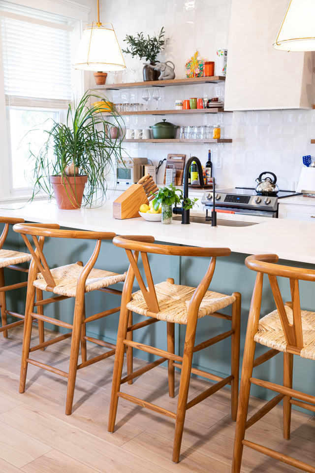 Four wooden counter stools with curved backs and woven natural fiber seats, lined up at a teal kitchen island surrounded by modern decor and potted plants