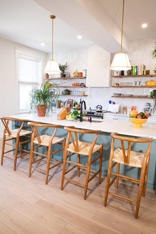 A bright kitchen featuring a teal island with four wooden counter stools with curved backs and woven seats, pendant lights, and open shelving with colorful dishes.