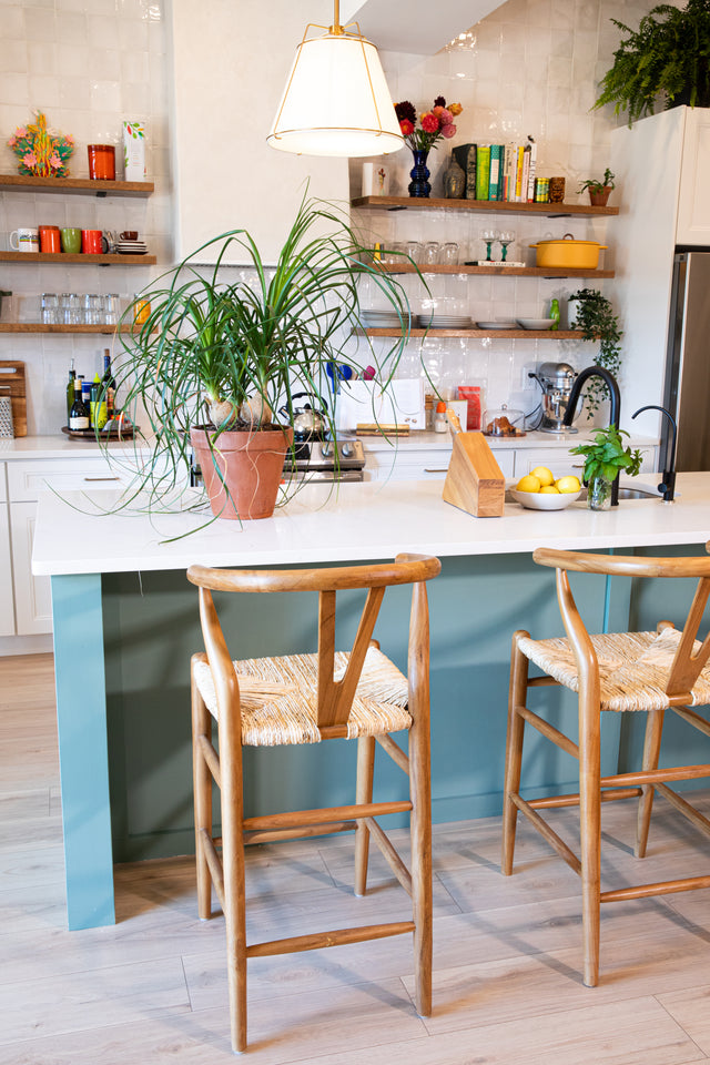 Two wooden counter stools with curved backs and woven seats positioned at a teal kitchen island, decorated with a large potted plant and modern kitchen accessories.