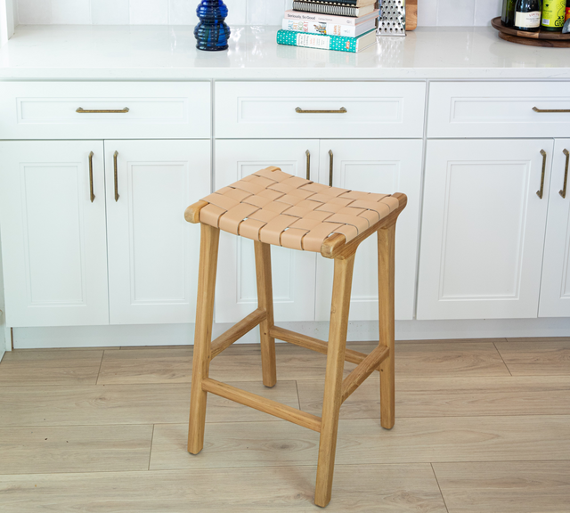 A wooden counter stool with a woven tan leather seat placed in front of white kitchen cabinets and brass hardware.