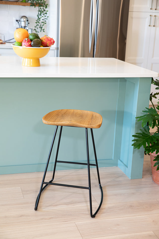 A counter stool with a contoured wooden seat and sleek black metal frame, positioned beside a teal kitchen island topped with a vibrant fruit bowl.